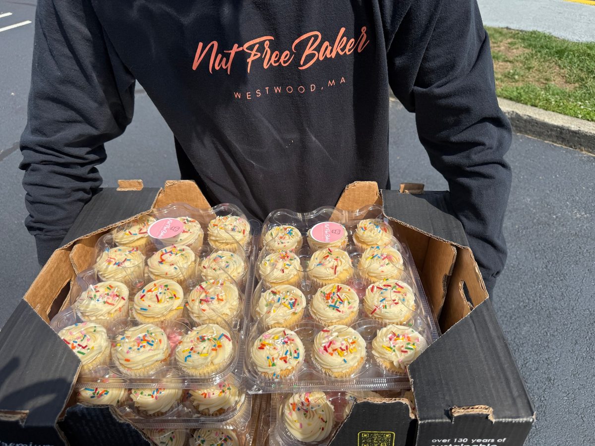 Trays of vanilla cupcakes with sprinkles, each pack labelled with a Nut Free Baker sticker, carried by someone in a Nut Free Baker hoodie