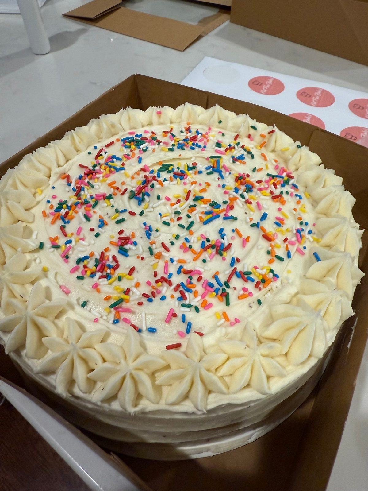 Vanilla cake with rainbow sprinkles and a piped border in a kraft box, next to a sheet of Nut Free Baker stickers